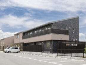 Two-story modern building with a dark stone lower facade, tan upper section, and a fenced entrance with a parked white van in front under a blue sky, likely a facility.