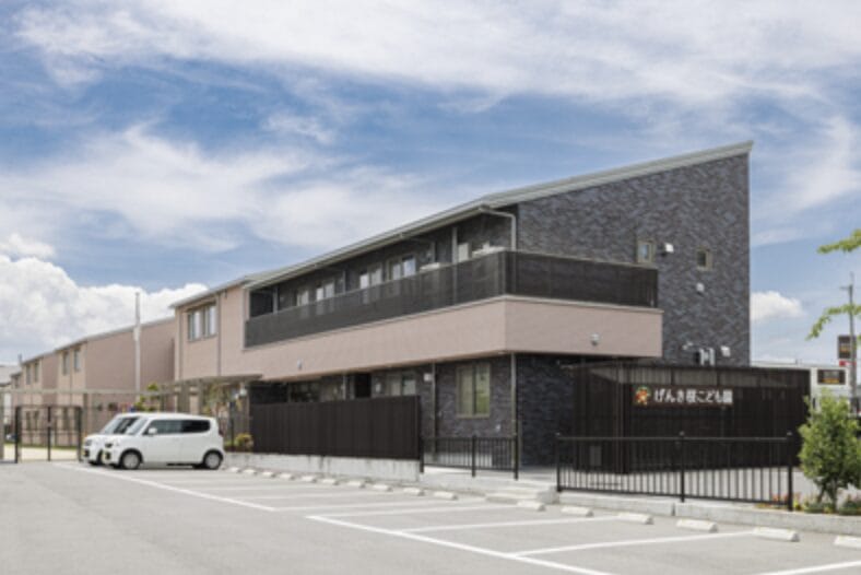 Two-story modern building with a dark stone lower facade, tan upper section, and a fenced entrance with a parked white van in front under a blue sky, likely a facility.