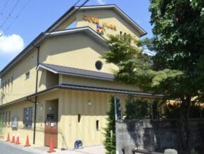 Front view of a yellow two-story childcare center with a sloped roof, orange traffic cones along the sidewalk, and a tree on the right.