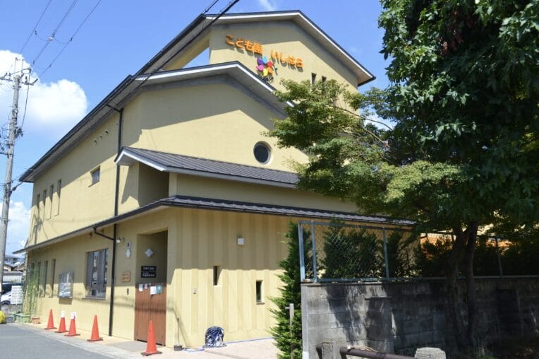 Front view of a yellow two-story childcare center with a sloped roof, orange traffic cones along the sidewalk, and a tree on the right.
