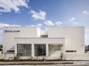 White modern office building with large glass storefronts and landscaped front yard under a blue sky, logo visible on the left side.