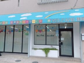 Storefront with a bright blue awning reading 'WELFARE' and Japanese text, large windows, and a scooter parked outside.