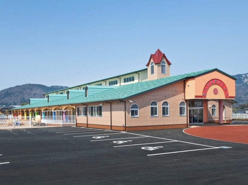 Brick school building with a green roof and red-topped tower, parking lot in the foreground, mountains in the background.