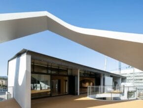 Modern building exterior with a large white curved canopy over a glass entrance on a sunny day.