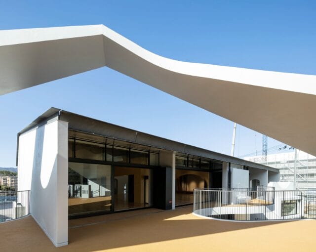 Modern building exterior with a large white curved canopy over a glass entrance on a sunny day.