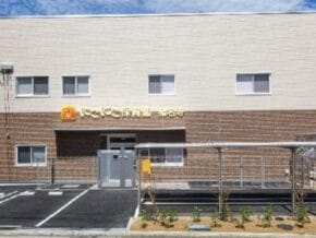 Two-tone building (beige upper, brown brick lower) with a sign in Japanese above the entrance and a parking area in front.