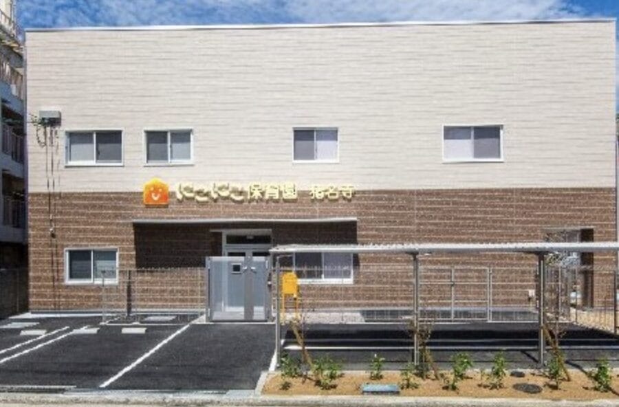 Two-tone building (beige upper, brown brick lower) with a sign in Japanese above the entrance and a parking area in front.