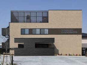 Facade of a modern beige and dark-gray building with a Japanese sign, likely a daycare center, and a parking area in front.