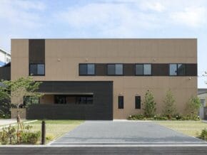 Two-story beige office building with a dark gray entrance extension, row of windows, and landscaped front yard.