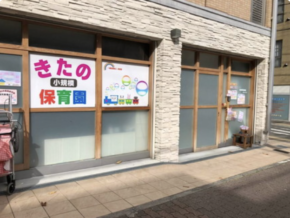 Exterior of a small Japanese daycare center with colorful signs in the windows, wooden-framed glass doors, and a sidewalk in front.