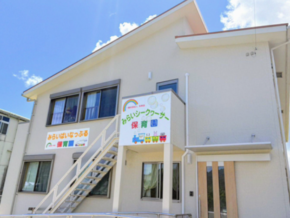 White two-story daycare/kindergarten building with an exterior staircase on the left, colorful Japanese signage, and a bright blue sky.