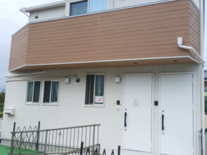 Two-story white brick building with a brown upper balcony, two white doors, and several windows; metal railing along a small ramp area.