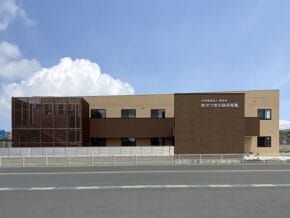 Two-story modern brown building with a darker central block and a vertical slat facade on the left, facing a paved street with a white fence in front.