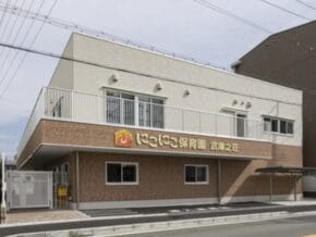 Two‑story daycare center with a tan brick lower level and light upper level, orange sign with a smiley logo, and a gated entrance facing the street.