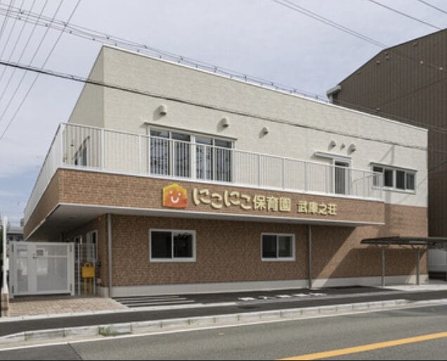 Screenshot | 保育士求人|大阪・兵庫・関西の保育士・幼稚園教諭の就職転職支援|保育のせかい Two‑story daycare center with a tan brick lower level and light upper level, orange sign with a smiley logo, and a gated entrance facing the street.