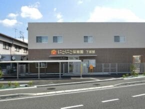Exterior view of a two-story daycare center with a covered entrance and a sign in Japanese (Niko Niko Hoikuen) on the front.