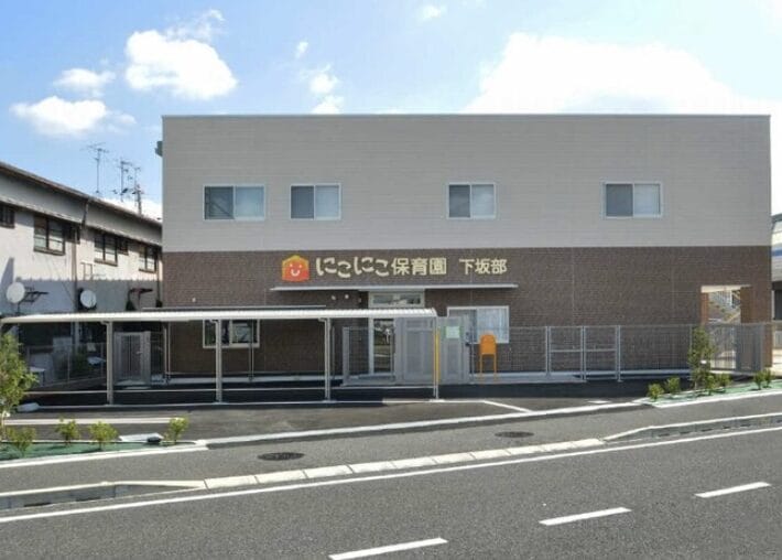 Exterior view of a two-story daycare center with a covered entrance and a sign in Japanese (Niko Niko Hoikuen) on the front.