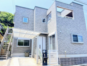Two-story grey brick house with a glass-covered entry, an external staircase on the left, a gated front entrance, and an upper-right balcony.
