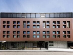 Modern red-brown office building with a grid of square windows and a low fence along the street side.