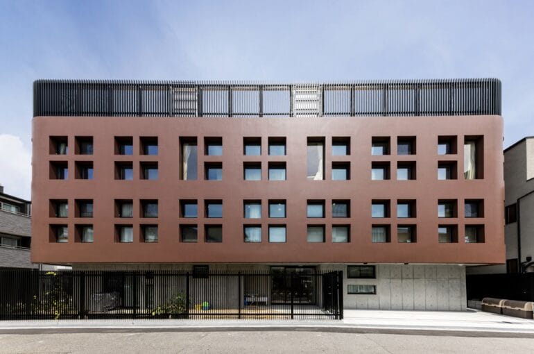 Modern red-brown office building with a grid of square windows and a low fence along the street side.