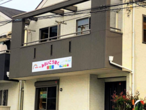 Two-story building with dark gray upper wall, beige lower wall, and a sign in Japanese above the entrance on a residential street.