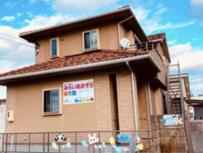 Two-story tan house with a red-tiled roof and a Japanese daycare sign, decorative animal cutouts along the fence in front, under a blue sky.