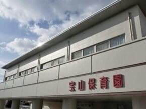 Two-story white daycare building with a red Japanese sign reading 宝山保育園 on the façade under a blue sky.