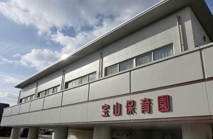 Two-story white daycare building with a red Japanese sign reading 宝山保育園 on the façade under a blue sky.