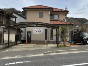 Two-story beige building with a red-tiled roof and curved translucent carport, a signboard at the front, orange traffic cones, and a van parked by the road.