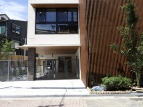 Modern townhouse with metal gate, brick pillar, and white façade; large windows above and wooden slat wall on the right, small trees and greenery nearby