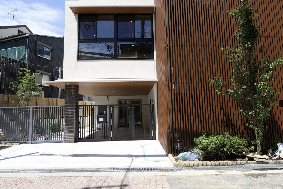 Modern townhouse with metal gate, brick pillar, and white façade; large windows above and wooden slat wall on the right, small trees and greenery nearby