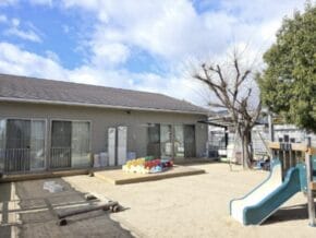A small outdoor playground in front of a single-story building with sliding glass doors, featuring a blue and white slide, colorful toy blocks, and bare trees under a sunny sky.
