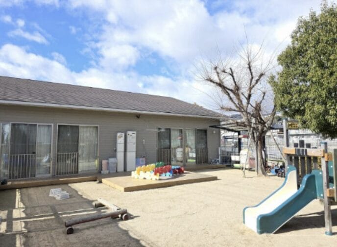 A small outdoor playground in front of a single-story building with sliding glass doors, featuring a blue and white slide, colorful toy blocks, and bare trees under a sunny sky.