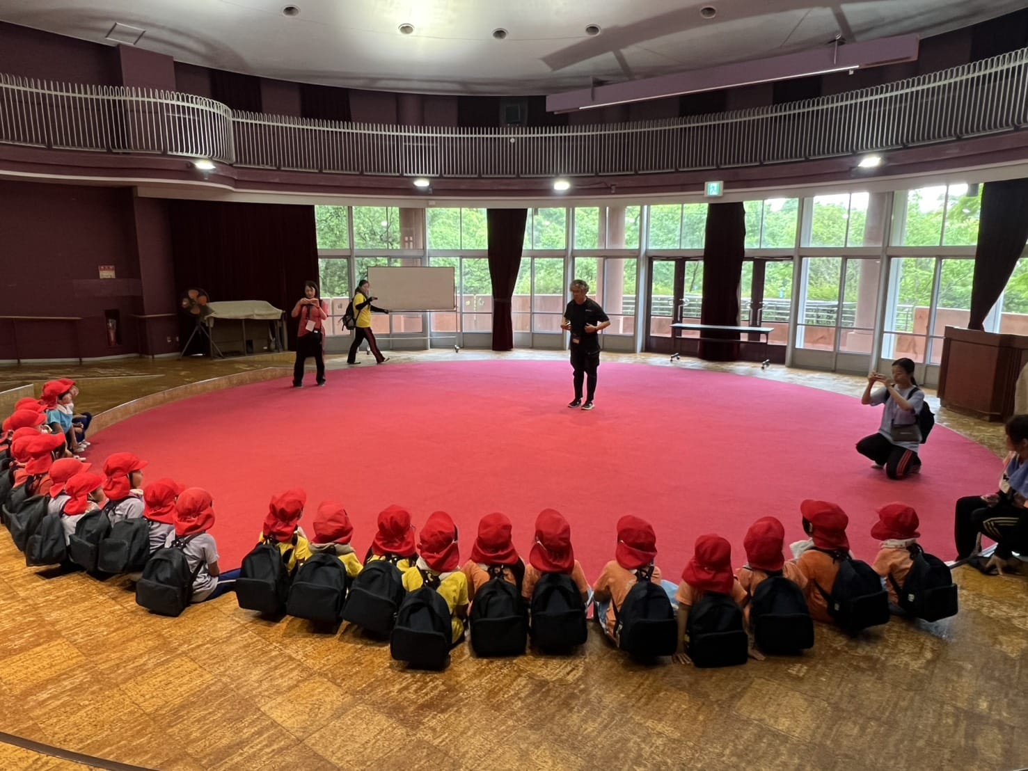 Children with red hats and backpacks sit in a circle on a red circular mat inside a large room as an instructor stands in the center.
