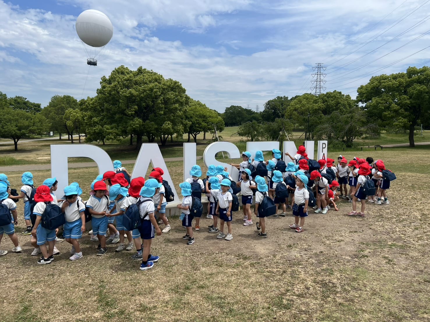 A line of young students in white shirts and blue shorts with backpacks, wearing red and light-blue hats, walking past large white letters in a park under a cloudy sky with a tethered balloon.