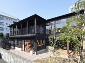 Two-story black wooden building with a long covered second‑floor balcony and large windows, surrounded by trees.