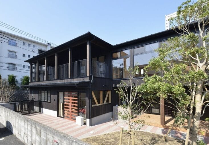 Two-story black wooden building with a long covered second‑floor balcony and large windows, surrounded by trees.