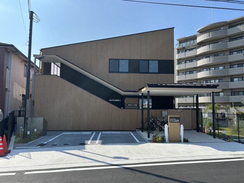 Brown modern building with a slanted roof, parking spaces in front, and bicycles under a canopy on the right.