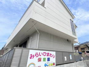 Exterior view of a modern white daycare center with a colorful sign reading 'みらいひまわり 保育園' and a metal fence, under a blue sky.
