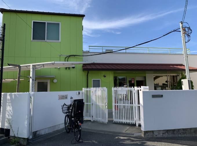 スクリーンショット 2026-10-28 162954 | 保育士求人|大阪・兵庫・関西の保育士・幼稚園教諭の就職転職支援|保育のせかい Two-story lime-green building with a white gate and a parked bicycle in front against a blue sky.