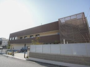 Two-story modern building with brown panels, a large metal lattice structure on the right, and a parked car in front.