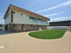 Two-story beige school building with a red tile roof, a sandy courtyard, and a circular grassy patch in front.