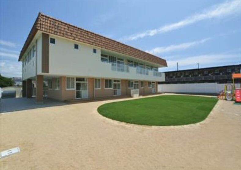 Two-story beige school building with a red tile roof, a sandy courtyard, and a circular grassy patch in front.