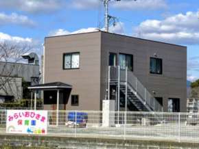 Two-story brown daycare building with an external metal staircase, fenced yard, and a colorful Japanese sign in front.