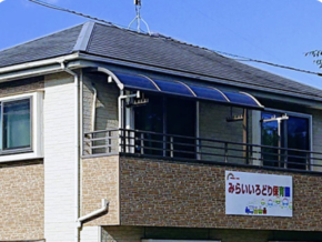 Two-story building with a blue curved awning over the upper‑story windows and a storefront sign in Japanese, indicating a daycare center.
