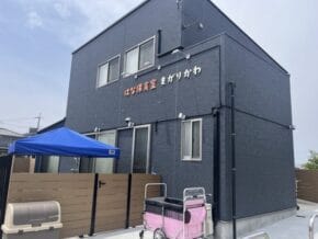 Exterior of a blue two-story daycare building with a Japanese sign, a blue canopy, and a pink stroller cart outside on a paved area.