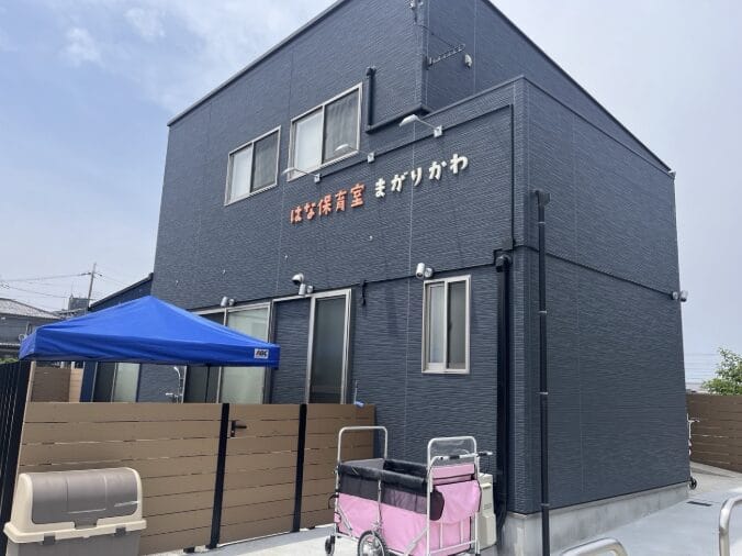 Exterior of a blue two-story daycare building with a Japanese sign, a blue canopy, and a pink stroller cart outside on a paved area.