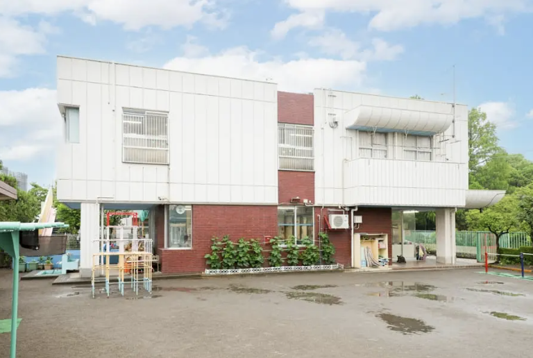 スクリーンショット 2026-09-11 161638 | 保育士求人|大阪・兵庫・関西の保育士・幼稚園教諭の就職転職支援|保育のせかい Two-story white-and-brick building with plants along the front and a small playground on the left on a wet paved area.