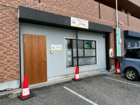Storefront entrance with a wooden door on the left, a large window in the middle, and a 'Sunny' sign above; orange traffic cones and a parked blue car nearby.