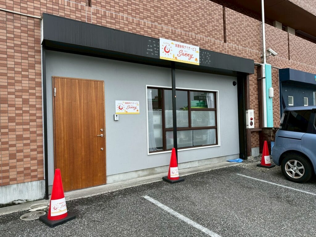Storefront entrance with a wooden door on the left, a large window in the middle, and a 'Sunny' sign above; orange traffic cones and a parked blue car nearby.